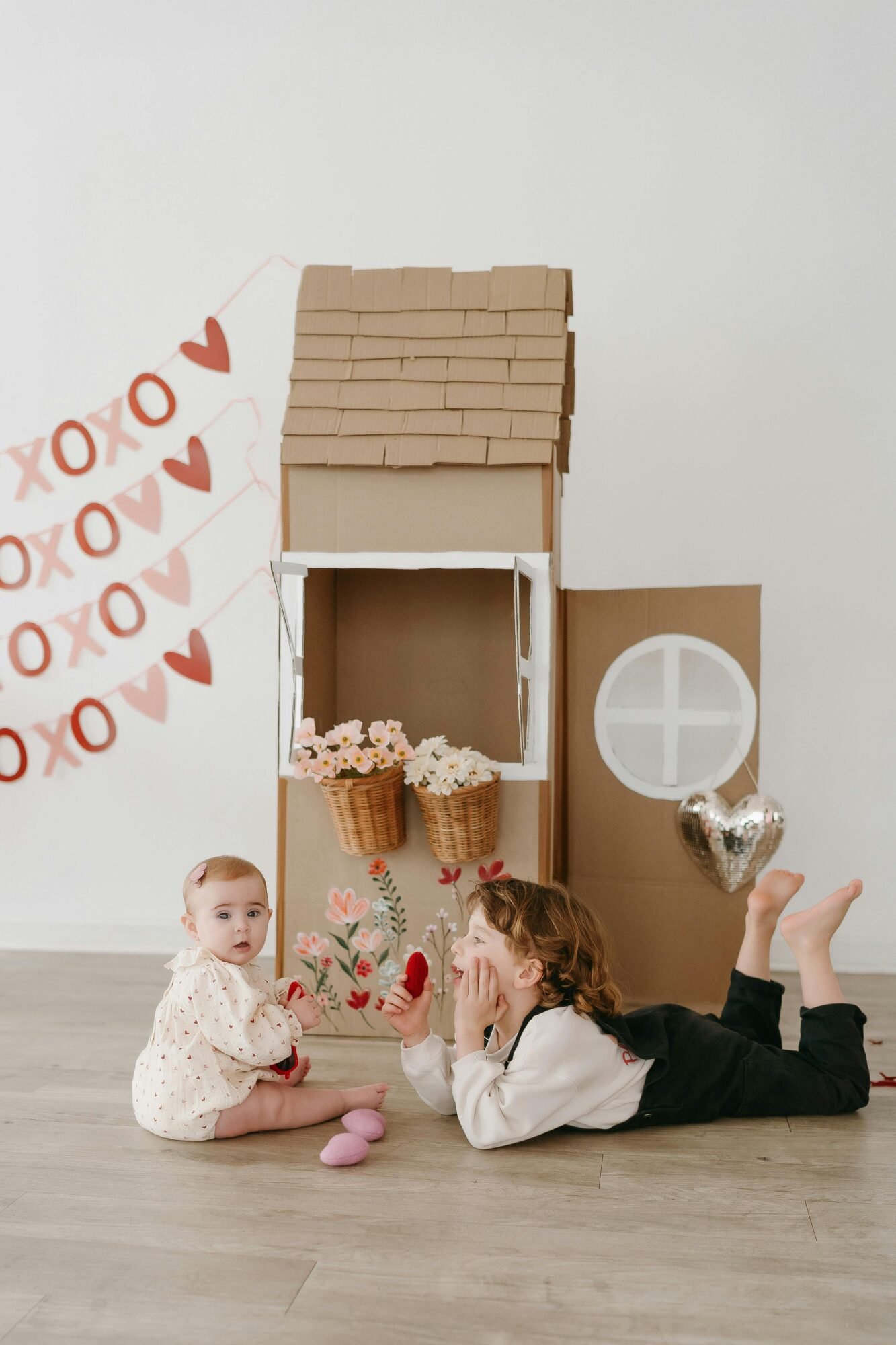 Two children playing with flowers in a charming cardboard house setup indoors.