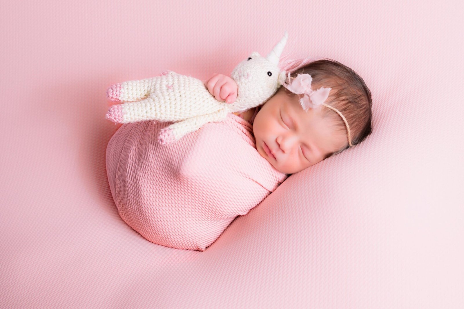Sweet newborn baby girl wrapped in pink with a unicorn toy on a soft background, exuding peace and innocence.